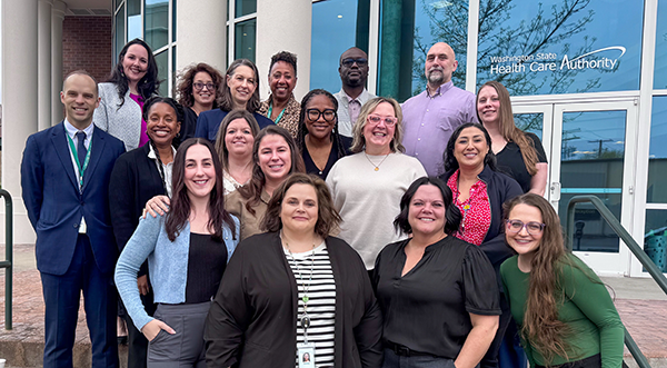team of prevention staff and SAMHSA officers standing in a group in front of a glass building with pillars in the background. People are staggered into four rows, smiling, and we wearing business casual clothing. It is bright yet overcast. 