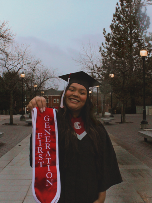 Woman smiling at camera wearing cap and gown from a graduation, holding up a swatch that says "First Generation."